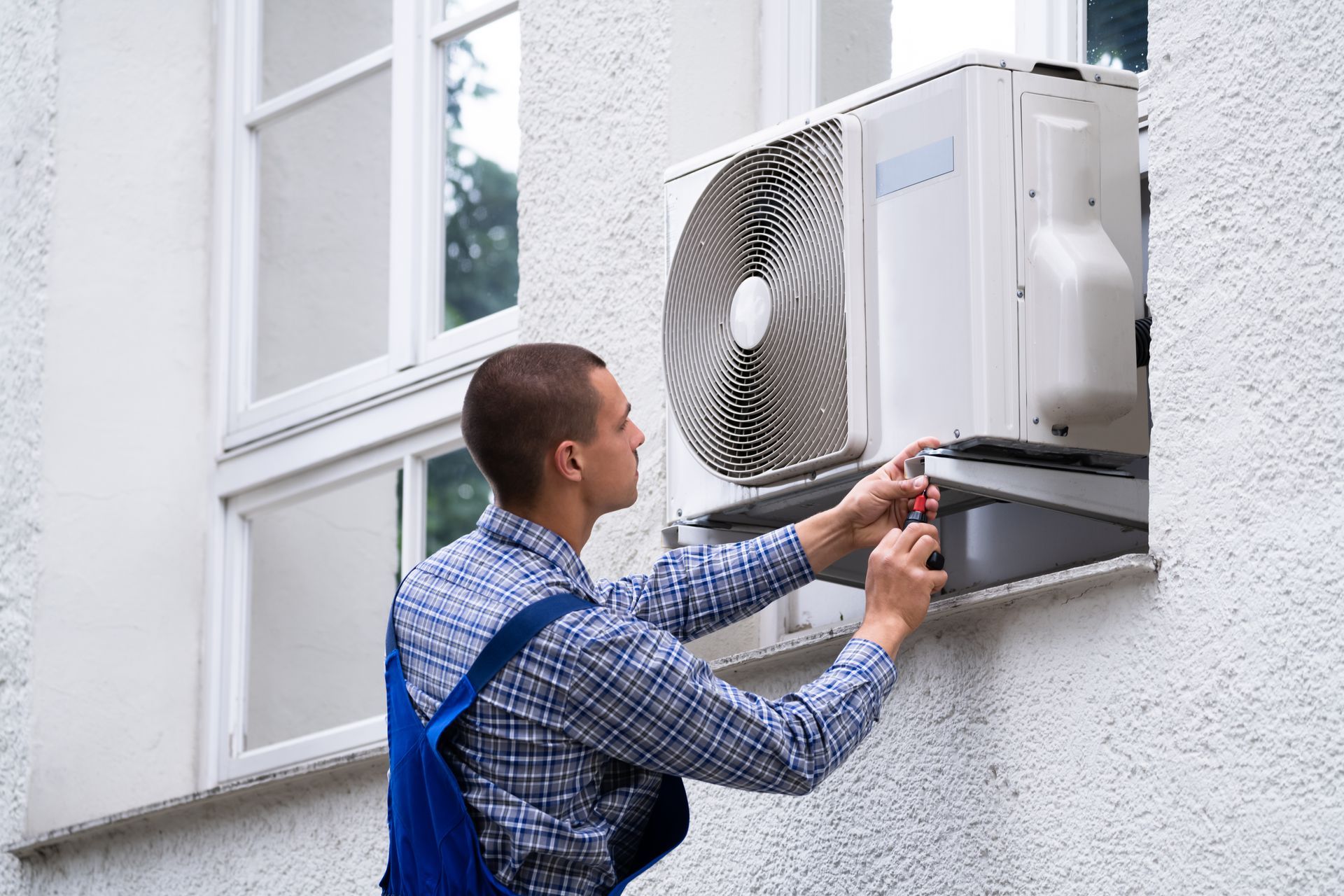 Technician servicing an outdoor air conditioner unit on a white building wall