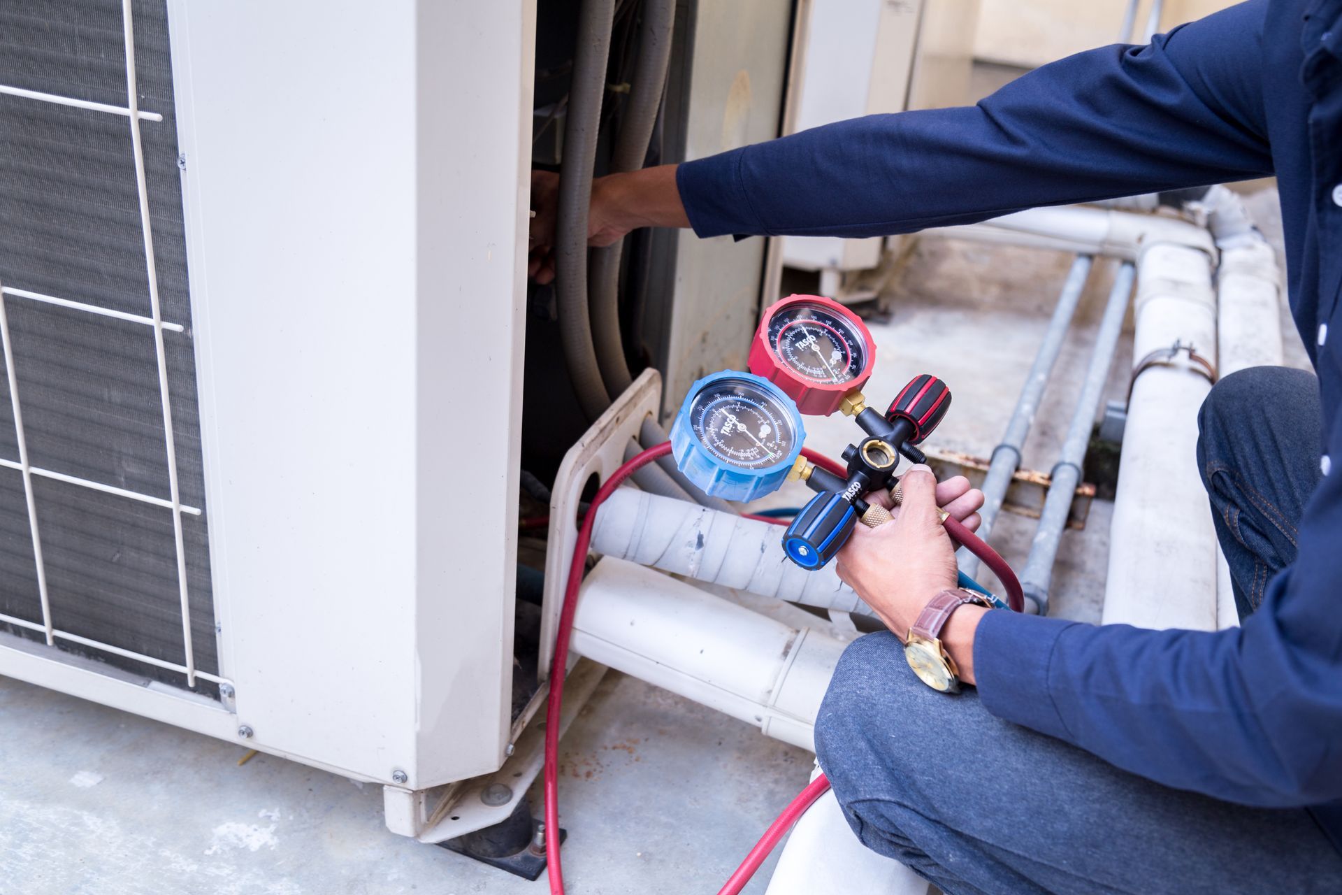 Technician using gauges to service an outdoor air-conditioning unit with red and blue hoses attached