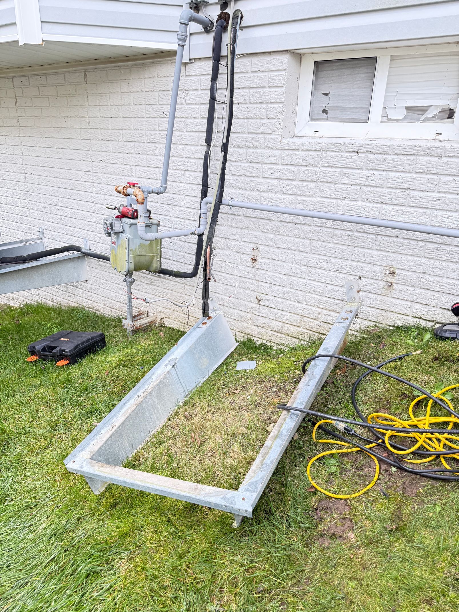 Metal gutter frame leaning against a white brick house beside a ladder, tools, and coiled yellow cord.