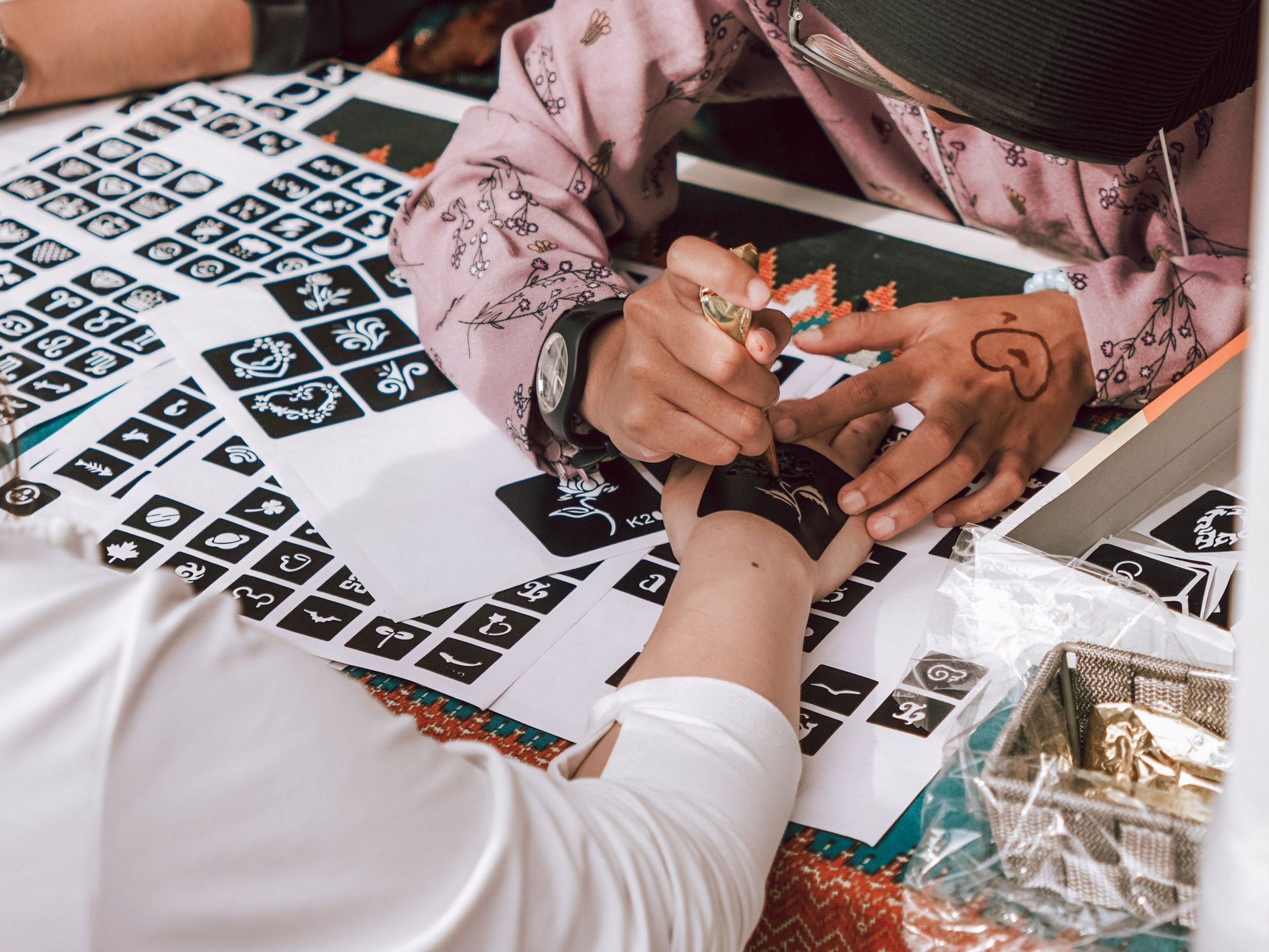 Applying henna to a person's hand at a decorated table with black-and-white pattern sheets.