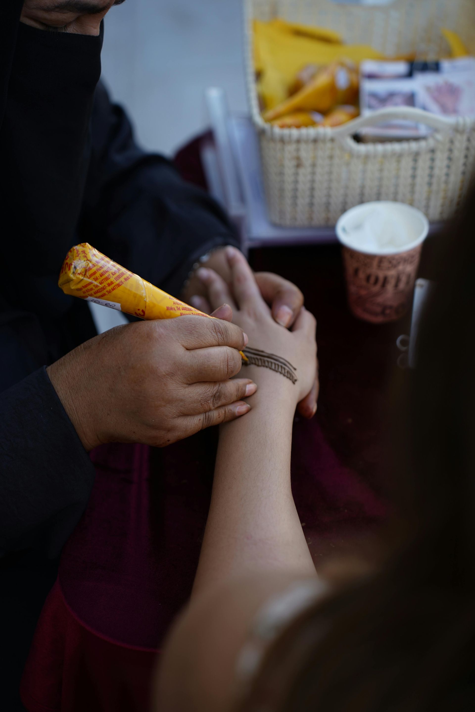 Hands painting a henna design on a wrist, with a cone applicator and paper cup nearby.