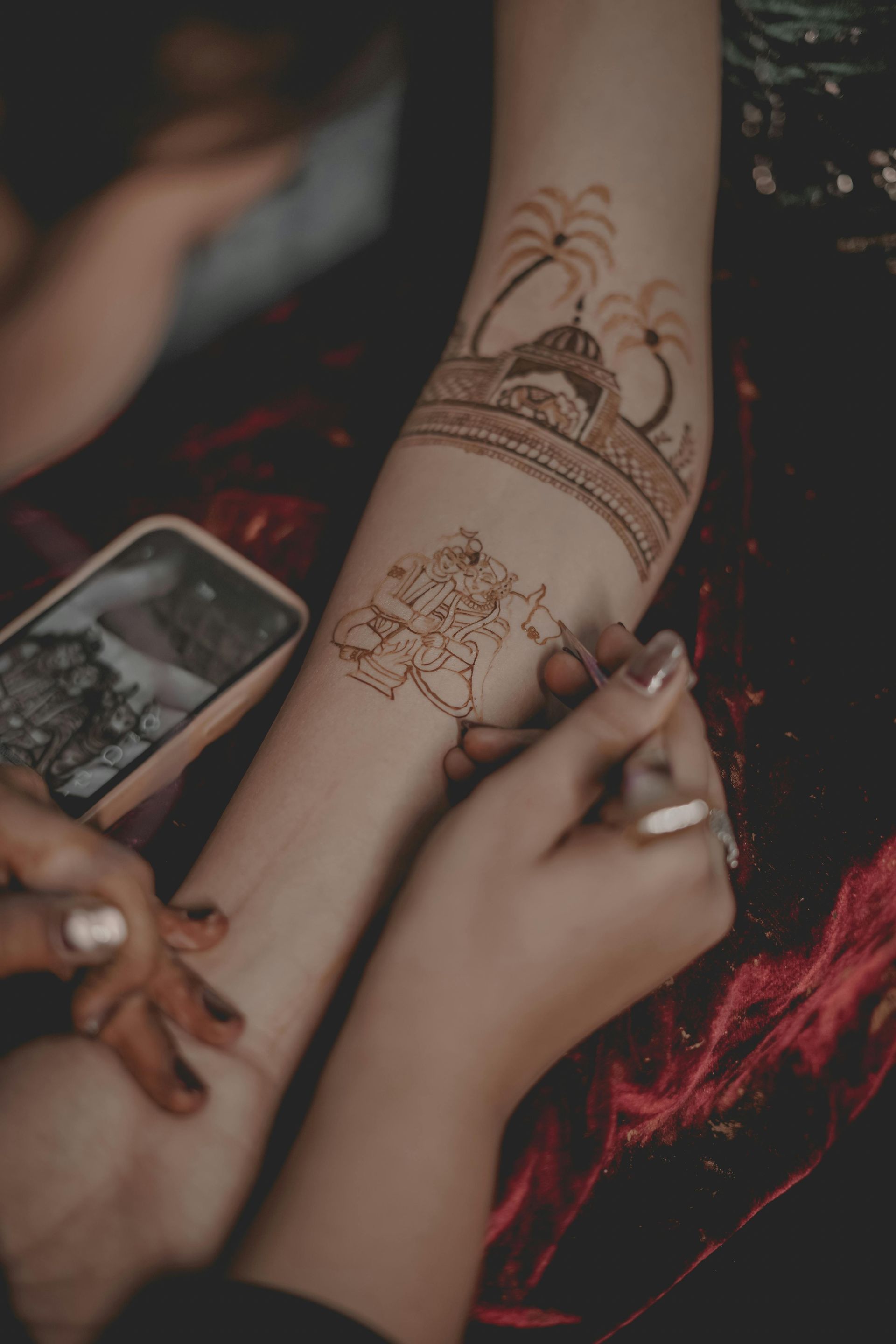 Hands applying henna to a forearm with intricate brown floral designs and bracelets.