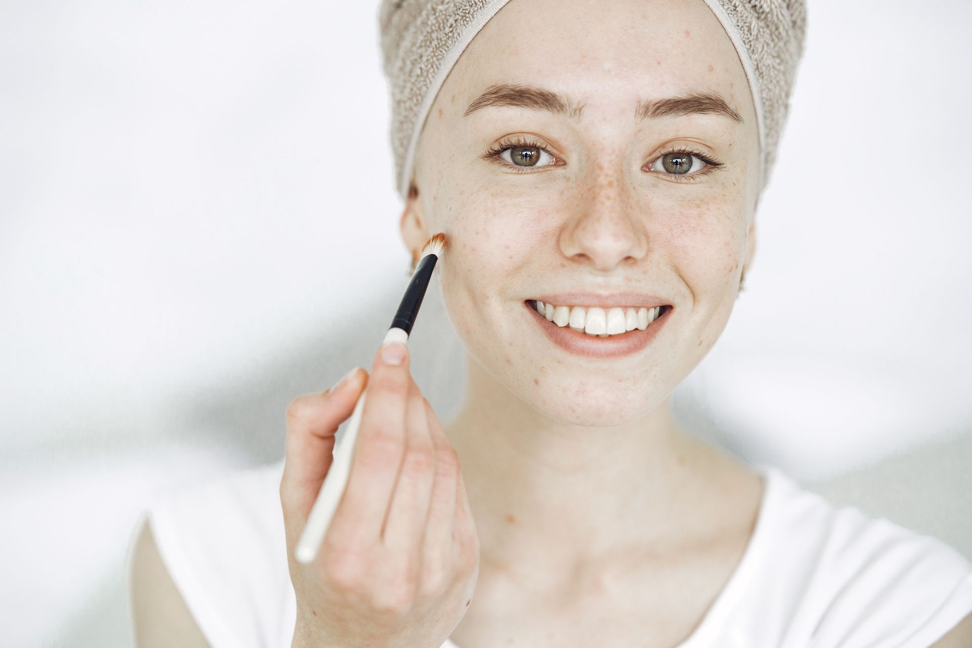 Woman applying makeup with a brush, smiling with a towel wrapped around her hair.