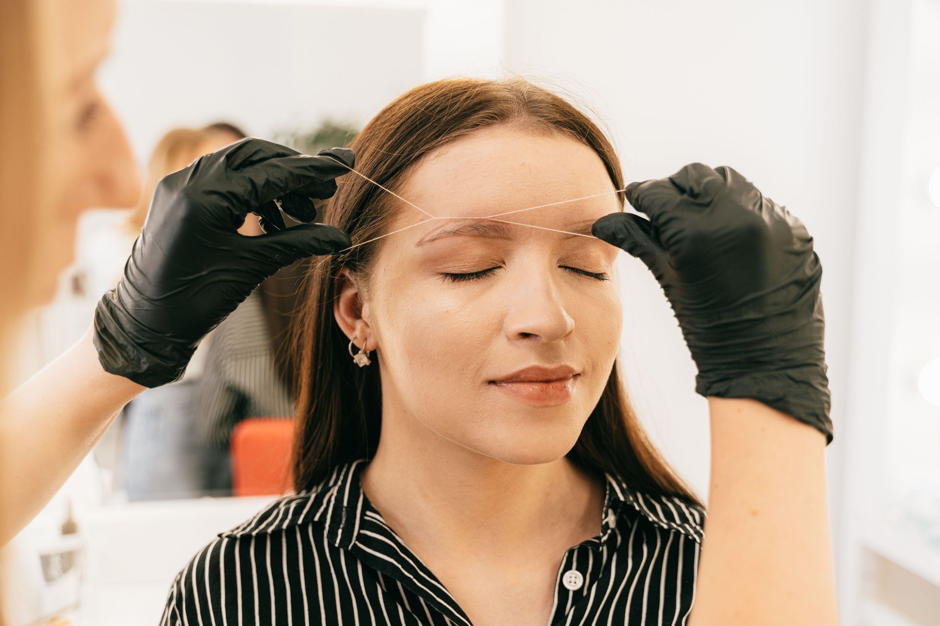Person receiving eyebrow treatment with gloved hands in a bright salon setting