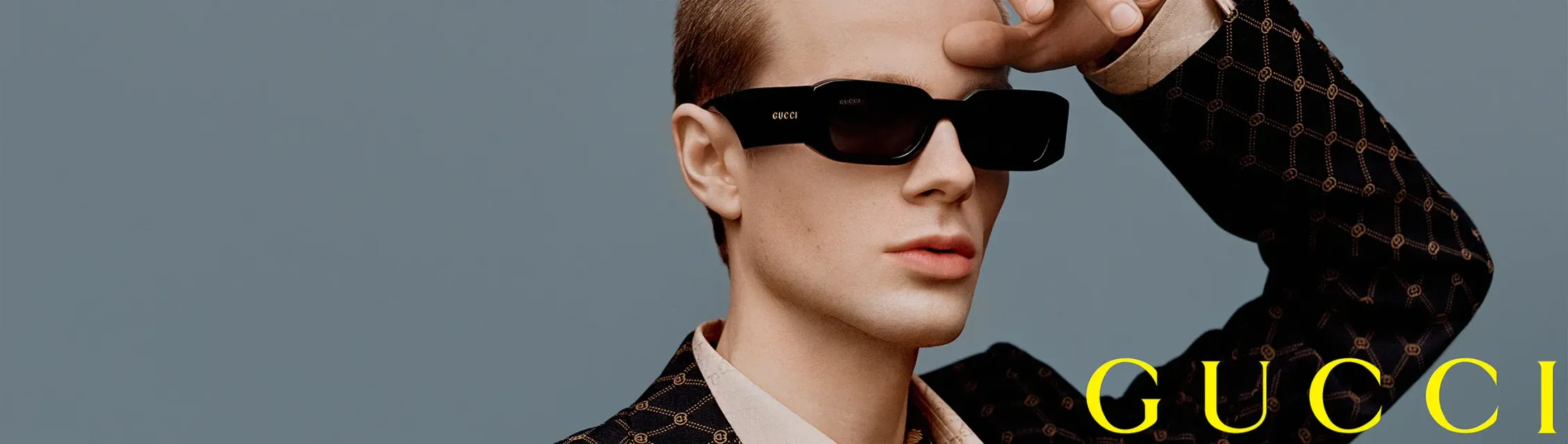 A man wearing sunglasses and a suit is covering his face with his hand.