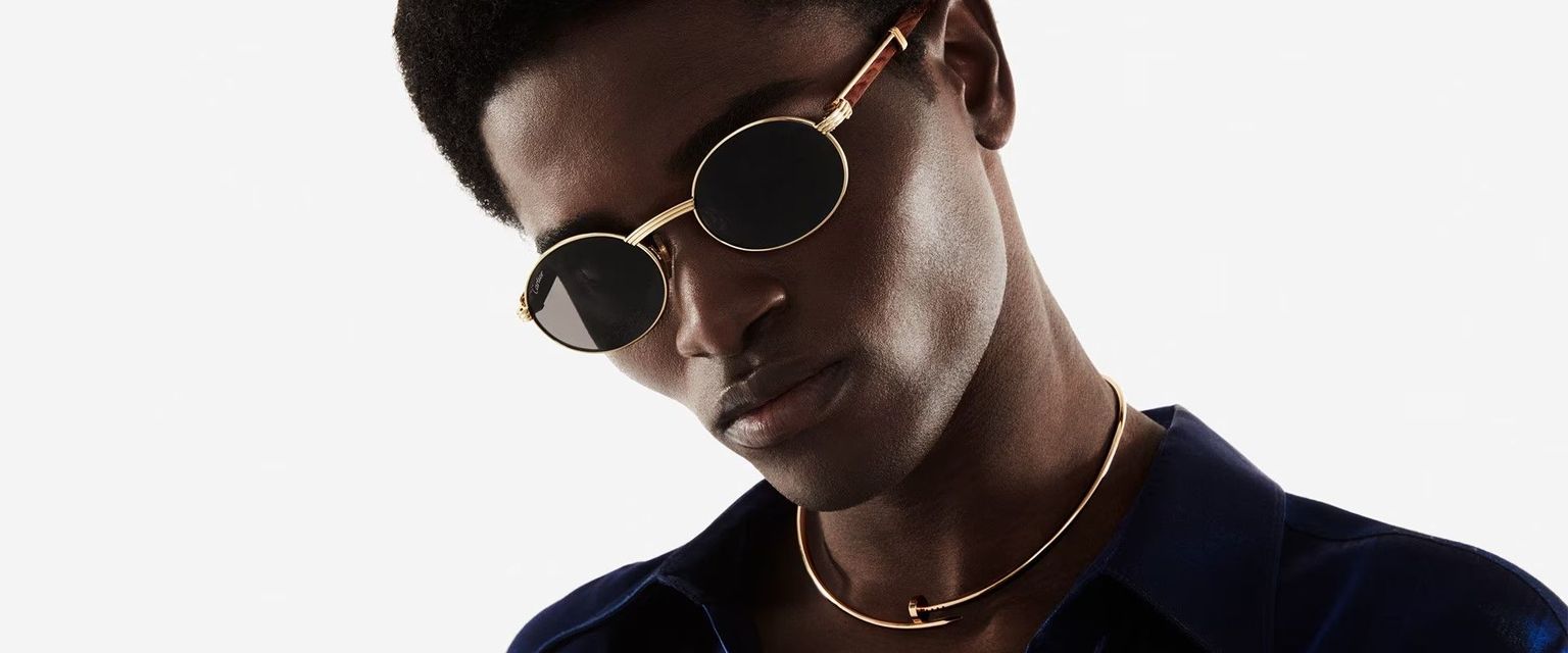 A man wearing sunglasses and a necklace is looking down.
