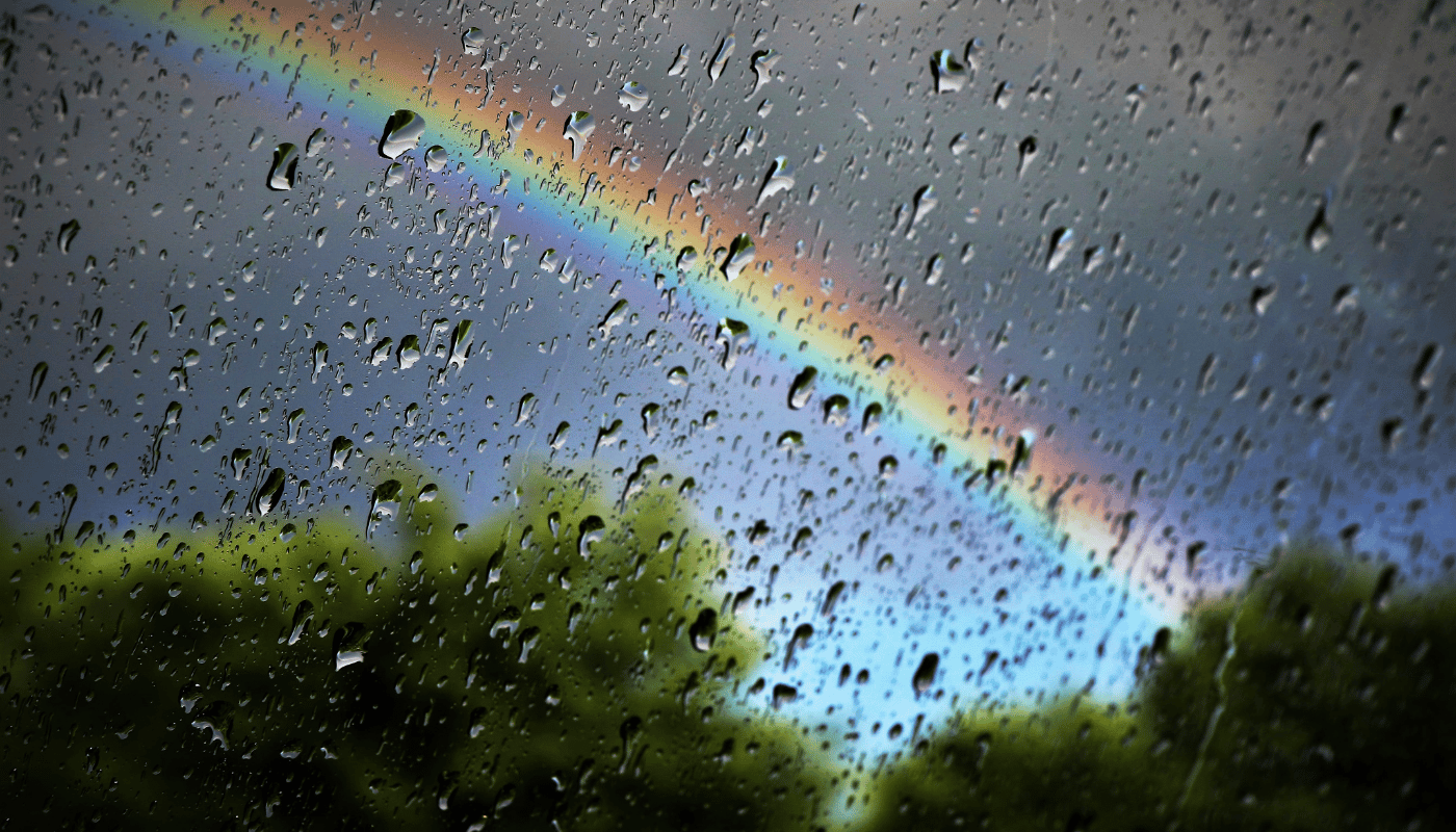 A rainbow is visible through the rain drops on a window.
