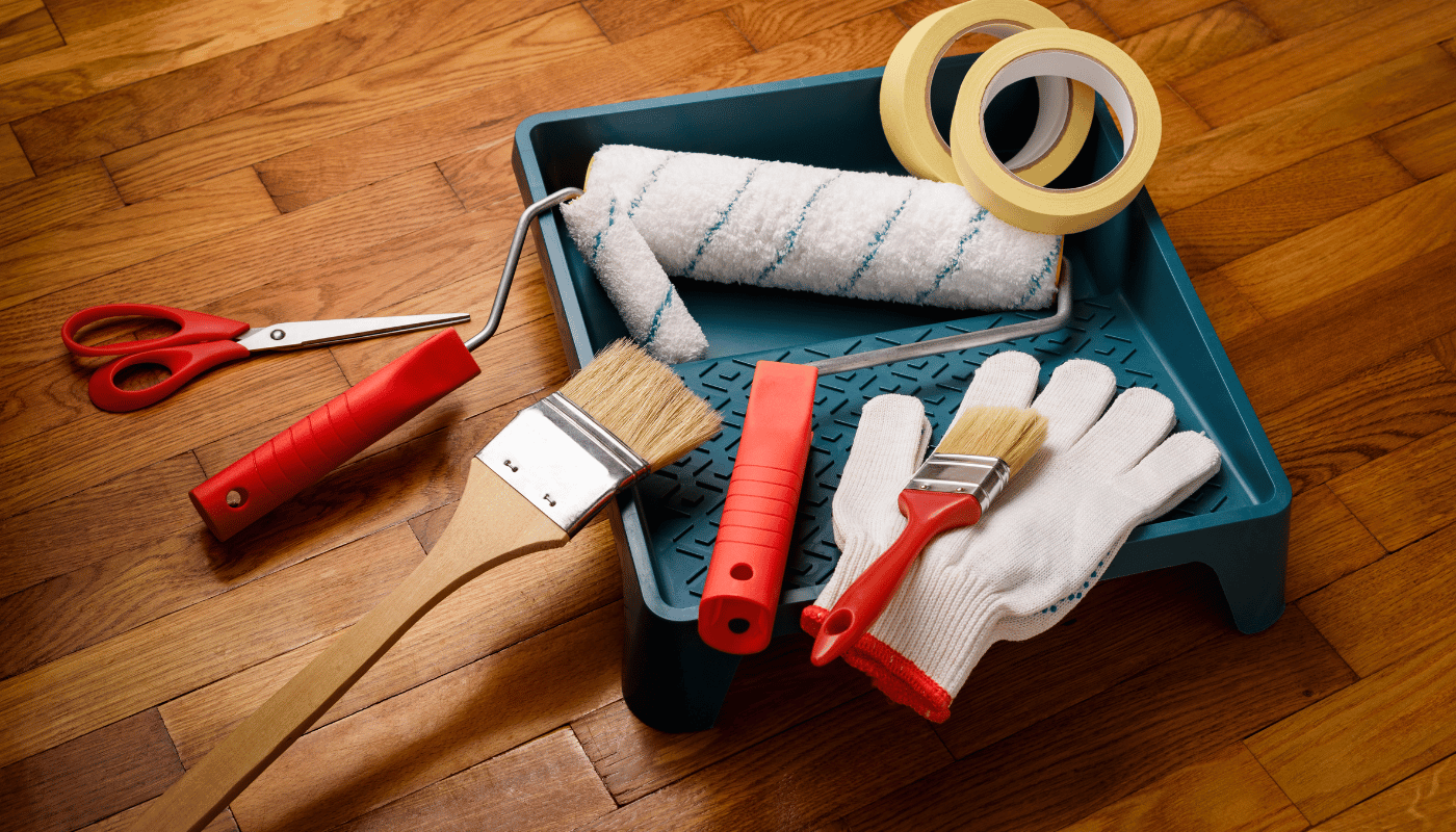 A tray with paint rollers , brushes , tape , scissors and gloves on a wooden floor.