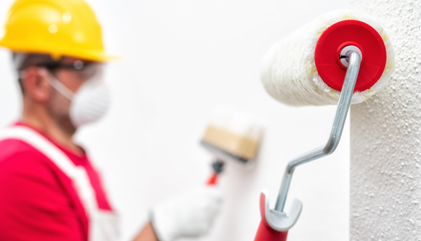 A man wearing a hard hat and a mask is painting a wall with a paint roller.