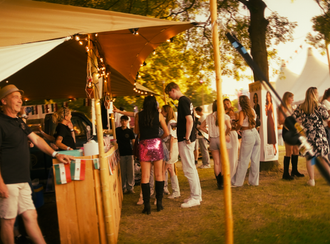Mensen staan ​​bij een eetkraam onder een beige luifel op een openluchtfestival tijdens een zonnige middag in het gouden uur.