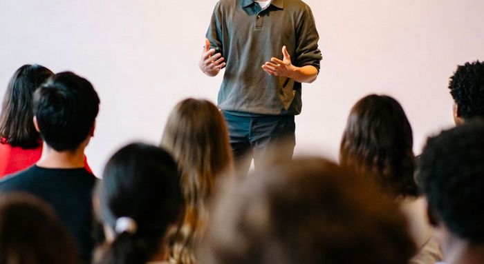 Man speaking to a group of people in a light-colored room. Audience is seated and facing the speaker.