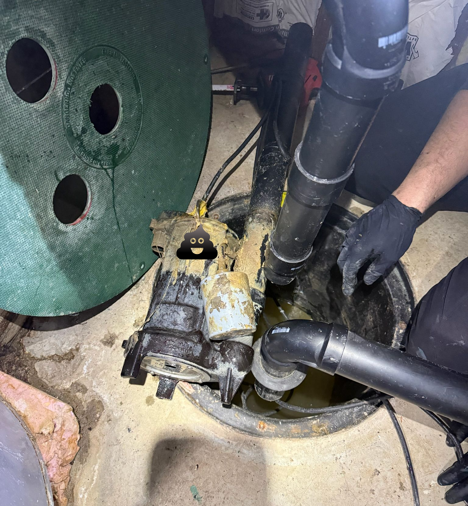 Sump pump in a pit; worker in black gloves inspects components. Black pipes, greenish lid, water present.
