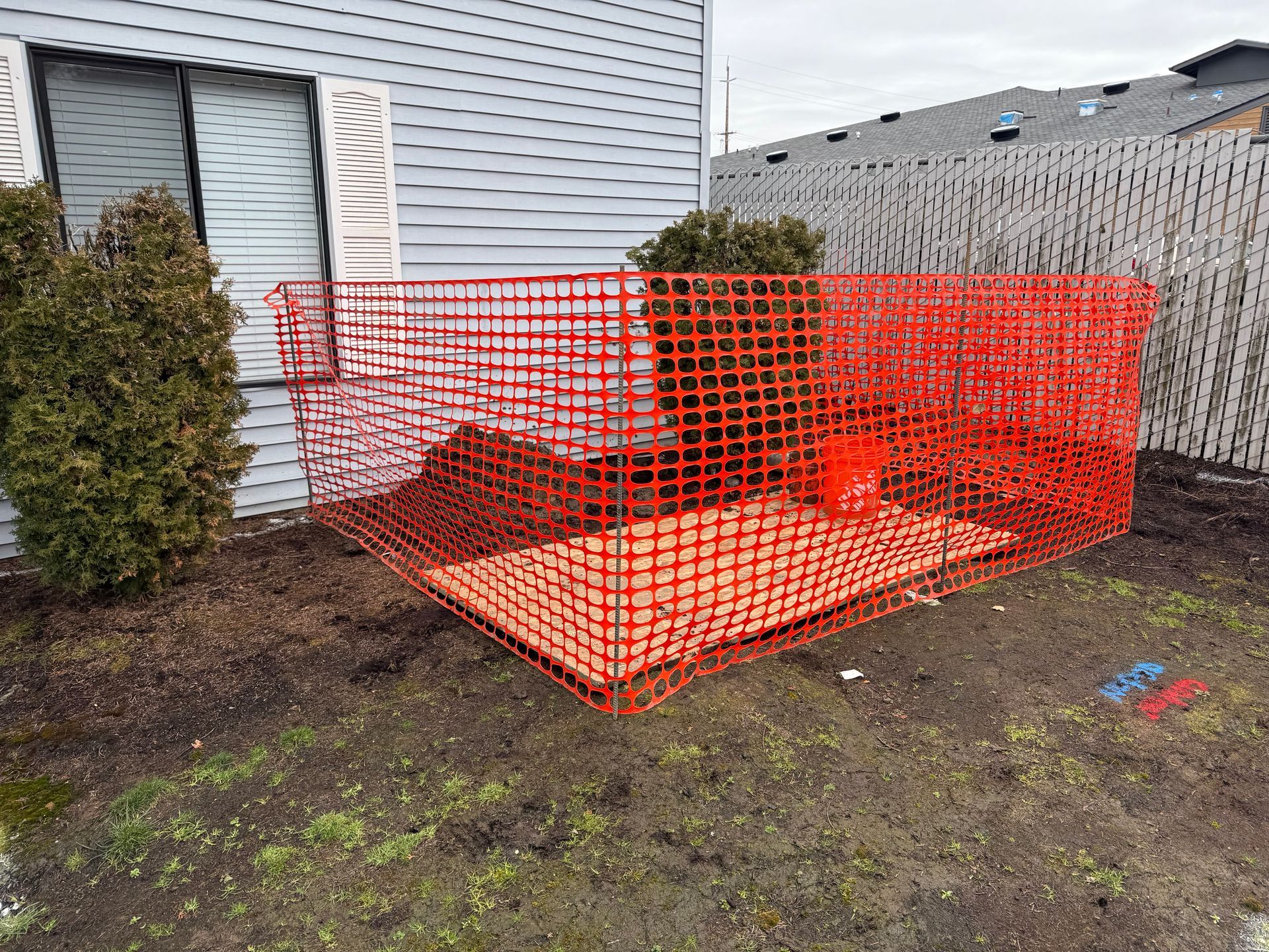Red construction fence encloses an area with a plant and debris outside a building.
