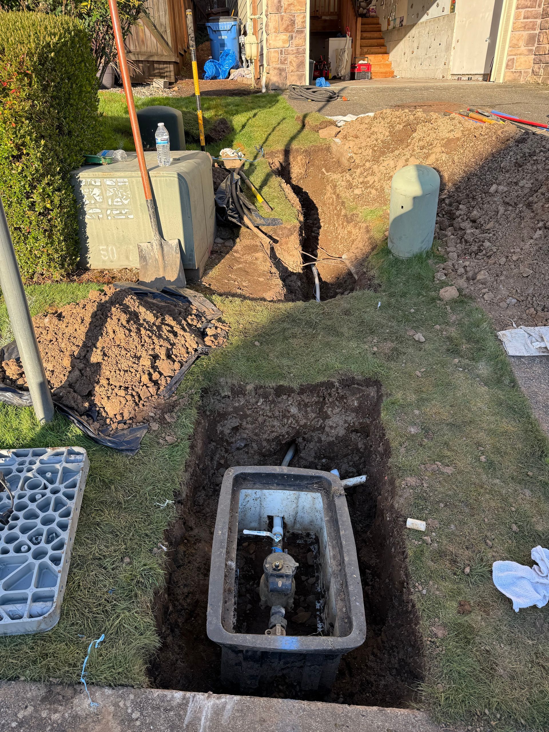 Backyard utility trench with exposed pipes and electrical boxes beside a small concrete access box