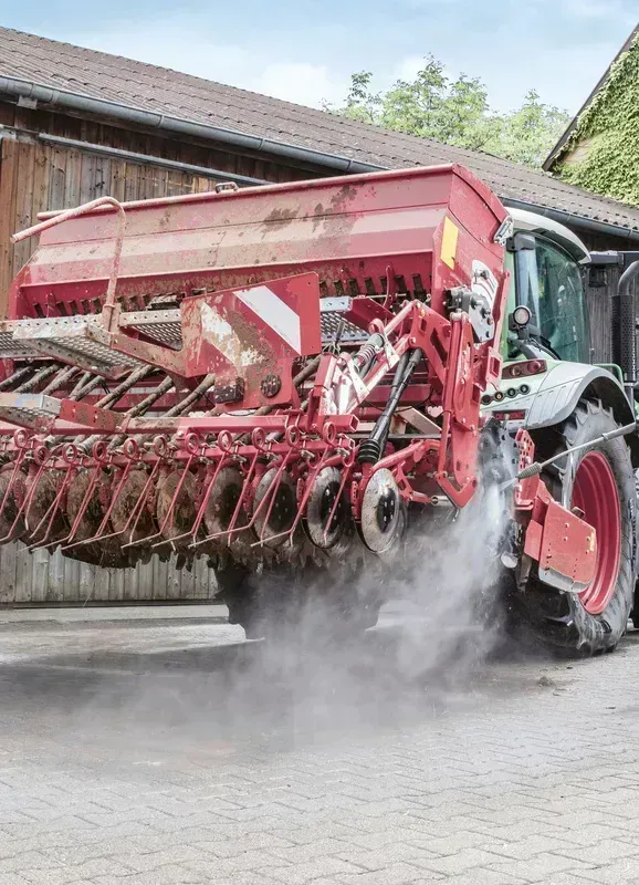 Red tractor with seeding equipment on a paved surface, kicking up dust. Barn in the background.