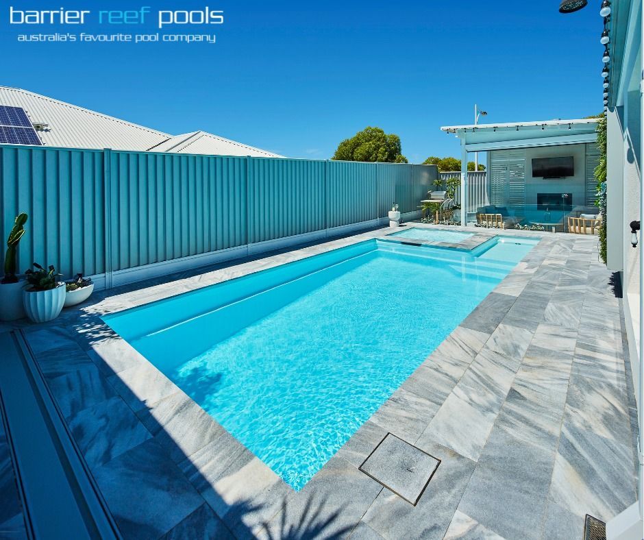 Swimming pool in a backyard with gray tile, a blue fence, and clear sky.