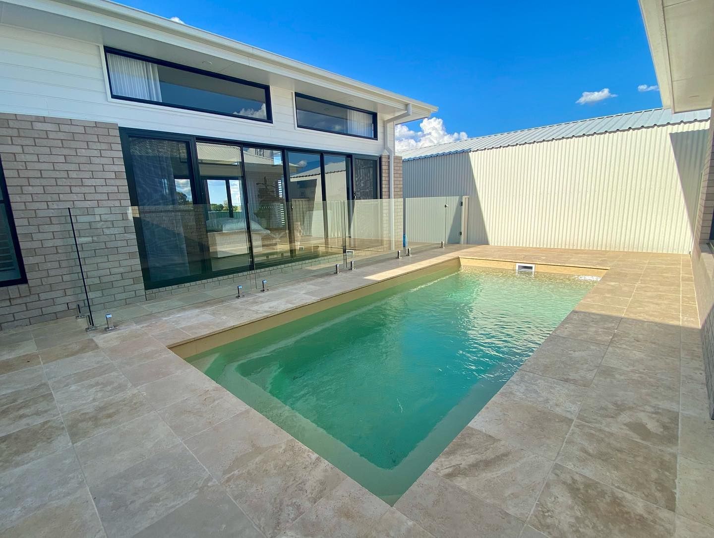 Backyard with rectangular pool; beige stone patio; modern house with glass doors. Sunny, blue sky.