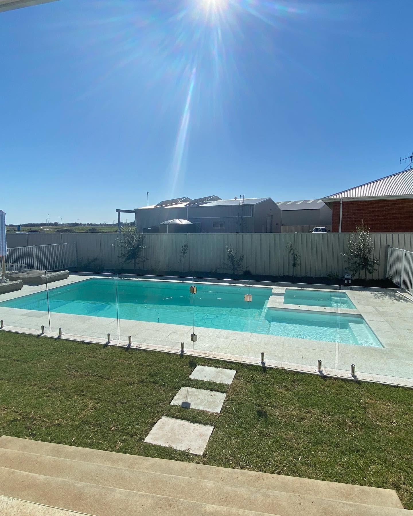 Backyard with a swimming pool, spa, and green lawn on a sunny day. White fence and blue sky in background.
