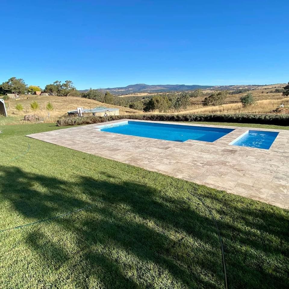 Rectangular pool and spa on stone patio, green lawn, rolling hills, clear blue sky.
