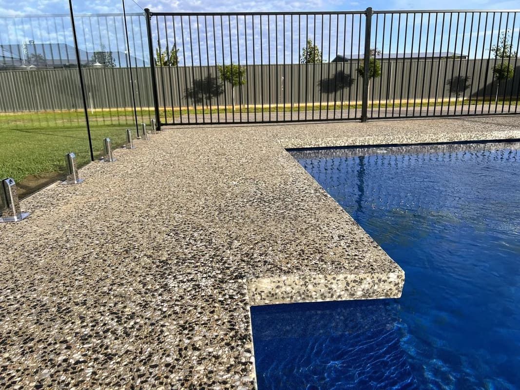 Poolside with textured concrete and blue water. Black fence and green lawn in background.