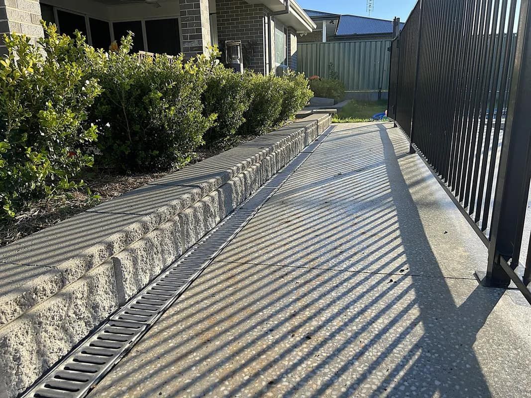 Concrete path with drainage, edged by a stone wall and black metal fence, casting shadows.
