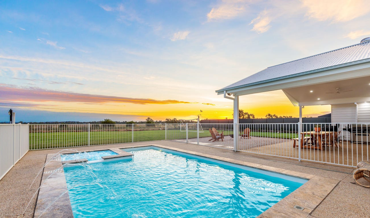 Pool next to a house with a white fence, overlooking a field under a sunset sky.