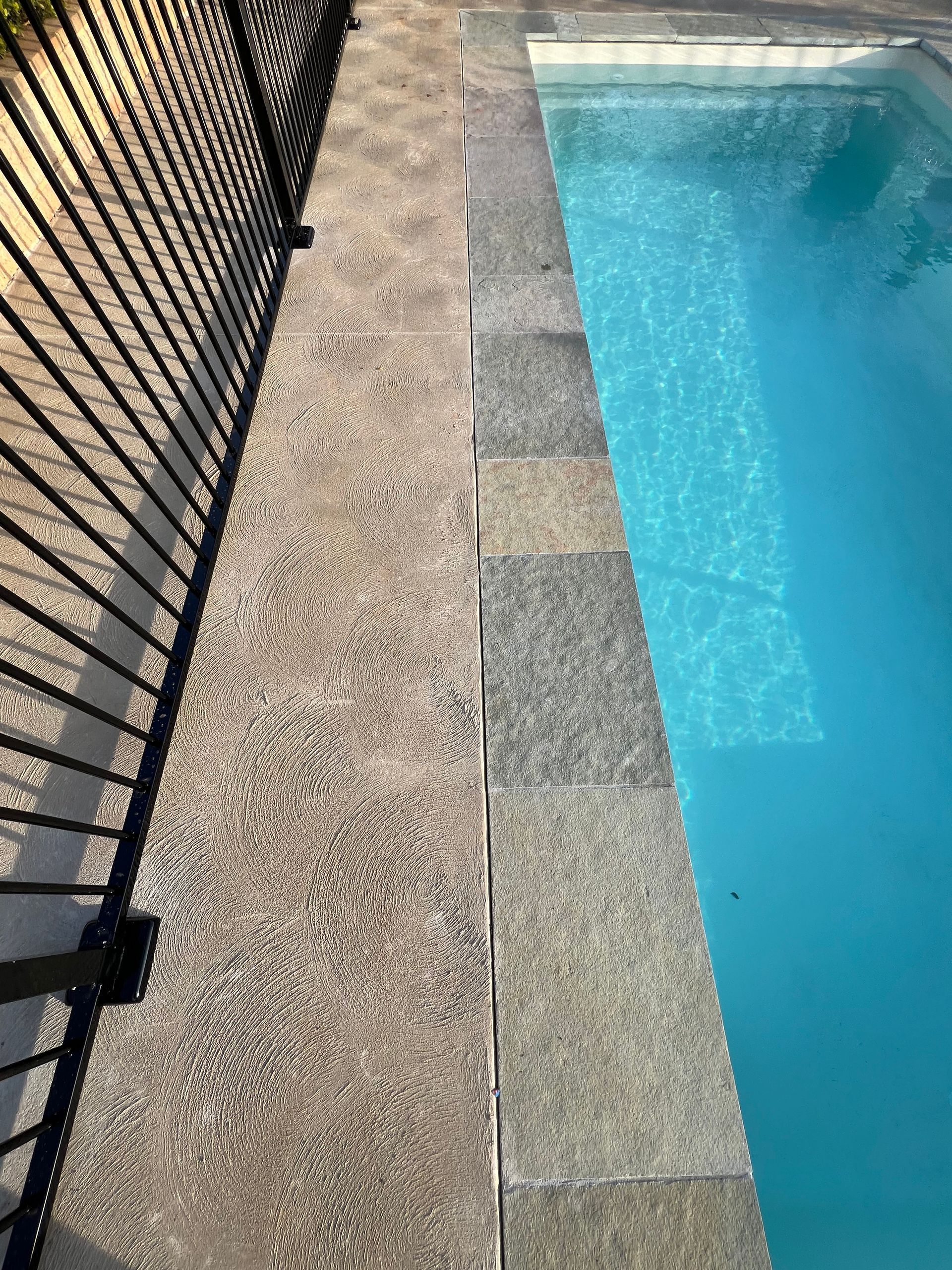 Poolside with a textured concrete deck, a black metal fence on the left, and a blue pool on the right.