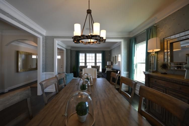 Long wooden dining table set for guests, chandelier, light green walls, windows in background.