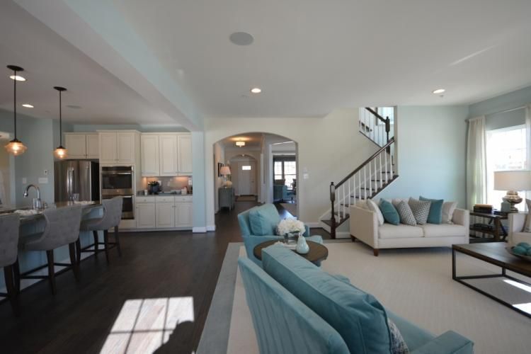 Open-concept living space with kitchen, living room, and staircase. Light blue walls, white cabinets, dark wood floors.
