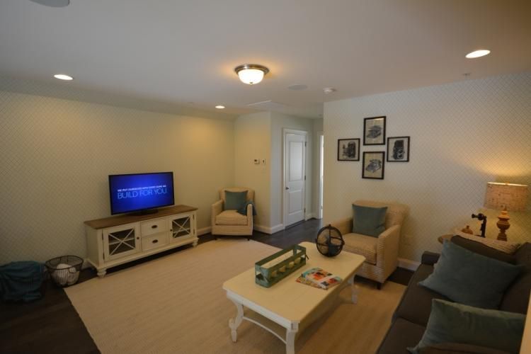 Living room with TV, beige chairs, and coffee table on a light rug. Photos on wall.