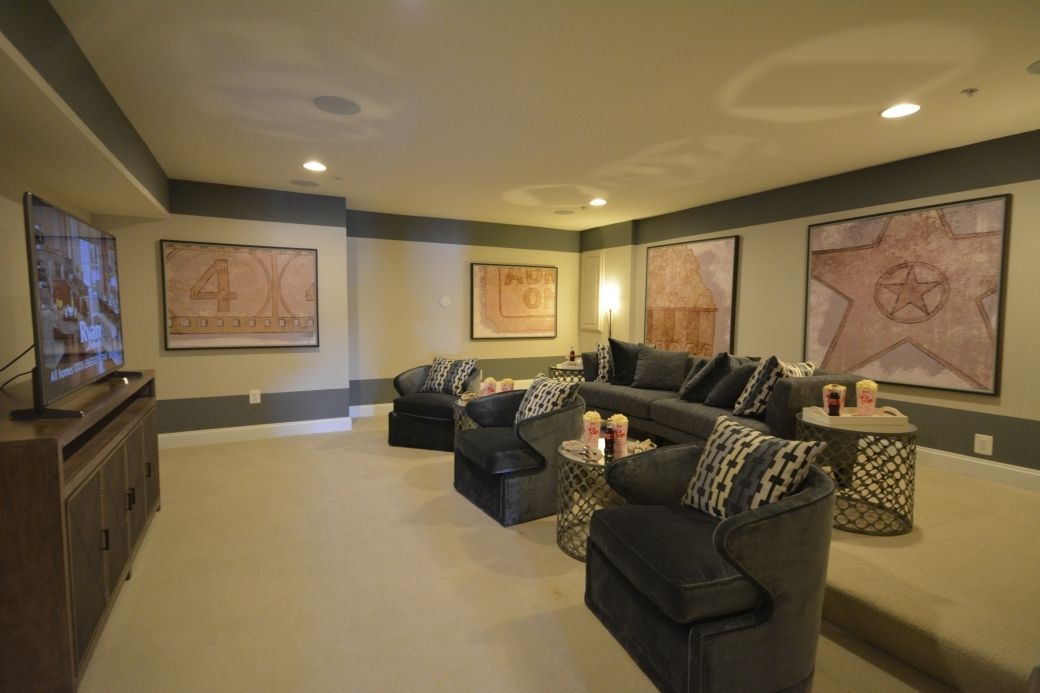 A modern home theater room with gray walls, sofa, chairs, framed art, and a large TV.