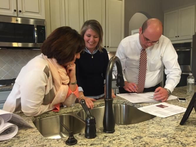 Three people reviewing blueprints in a kitchen with granite countertops, stainless steel sink, and dark faucet.