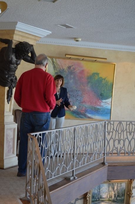 Man and woman on balcony looking at artwork. The man wears a red sweater and jeans. The woman wears a blazer.