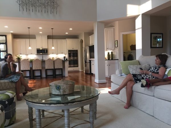 Two women relax in a spacious living room with a view into the kitchen, light wood floors, and neutral tones.