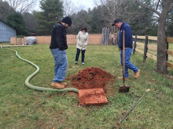 Three people inspecting an open septic tank in a yard with a green hose, shovel, and fence.