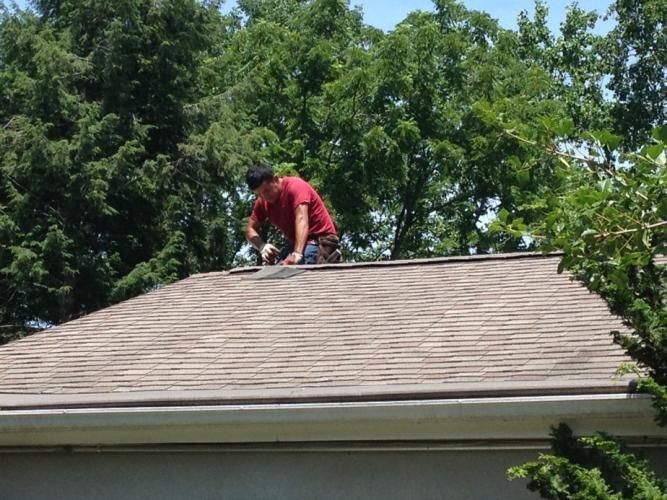 Man in red shirt on a roof, working on shingles. Trees in the background, sunny day.