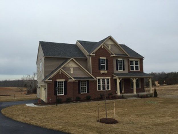 Two-story house with brick and tan siding, black shutters, and a porch, set in a grassy yard under an overcast sky.