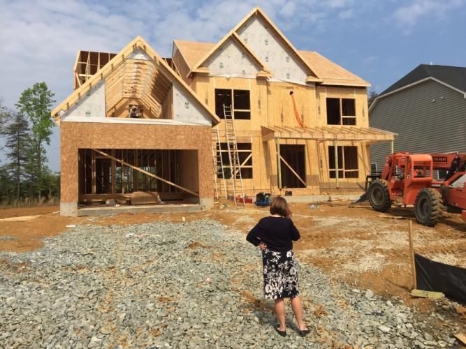 Woman standing near a house under construction; orange machinery on the right, cloudy sky overhead.