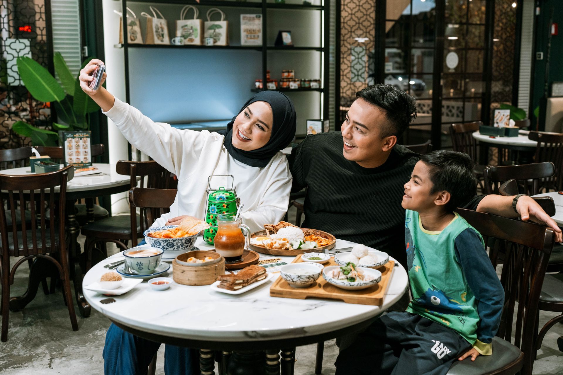 A happy Malay family sitting at a dining table, smiling and taking a selfie together with their smartphone
