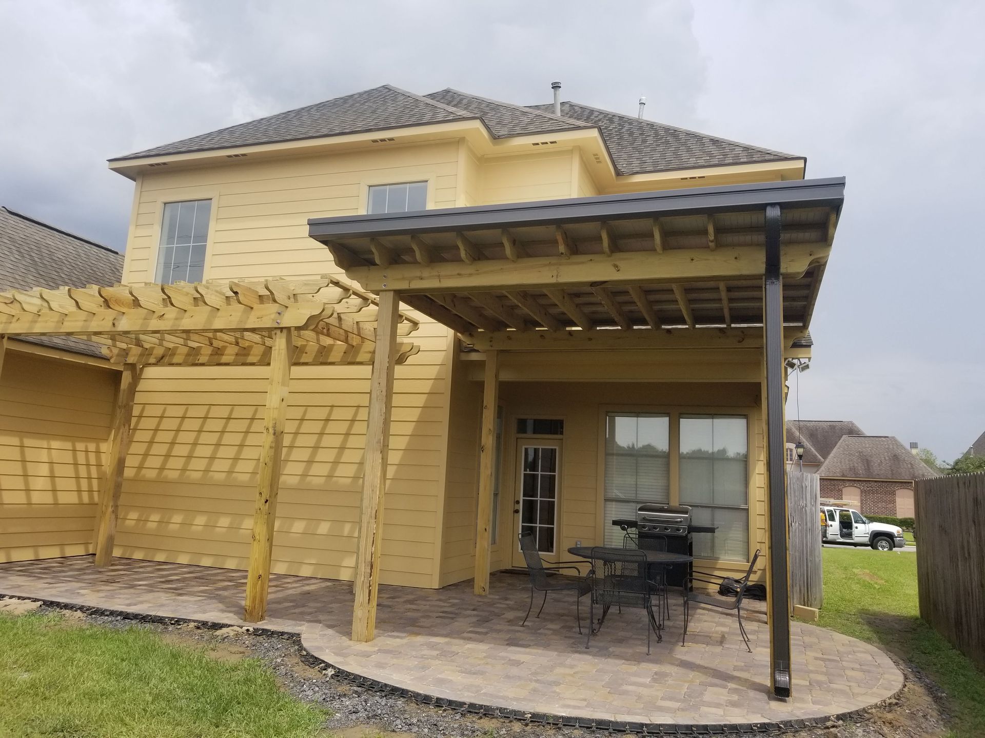 Yellow two-story house with a covered patio, wooden pergola, and paved backyard seating area