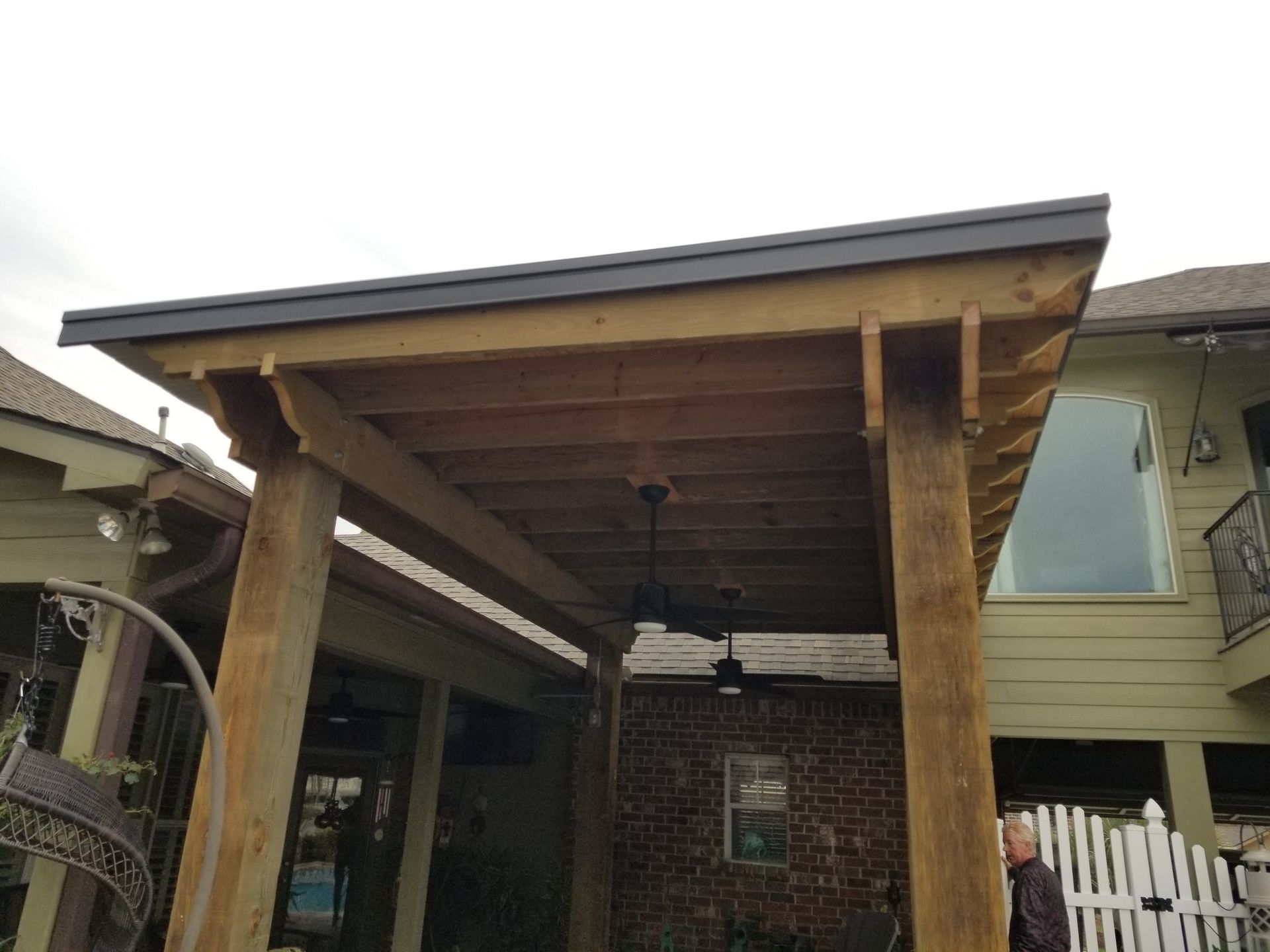 Covered patio with wood posts and dark roof beside a brick house, with a white fence in the background