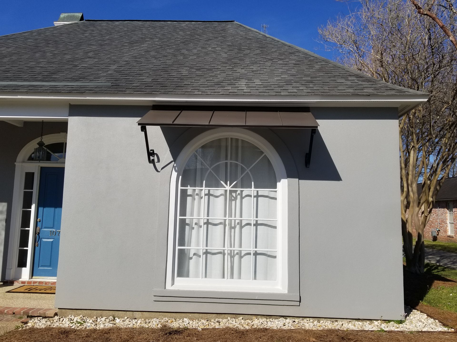 Gray house exterior with arched window, blue front door, and dark shingled roof in daylight