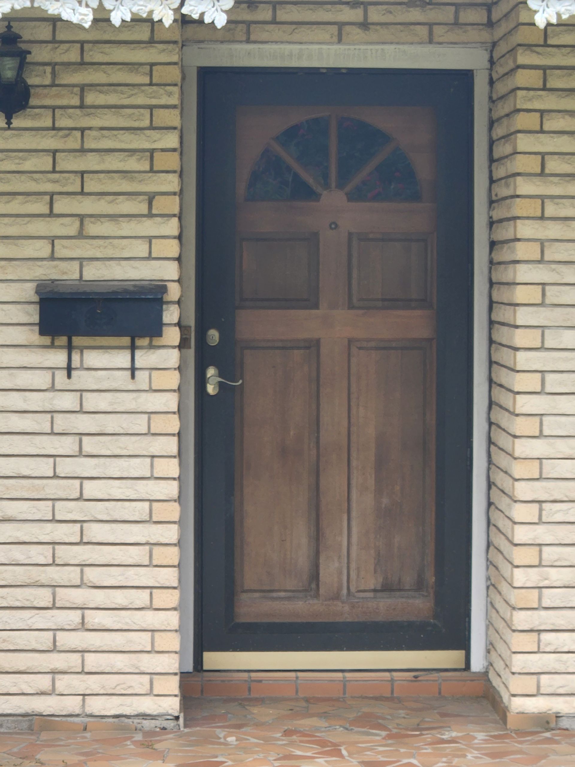 Wooden front door with arched window in a brick exterior wall, beside a black mailbox.
