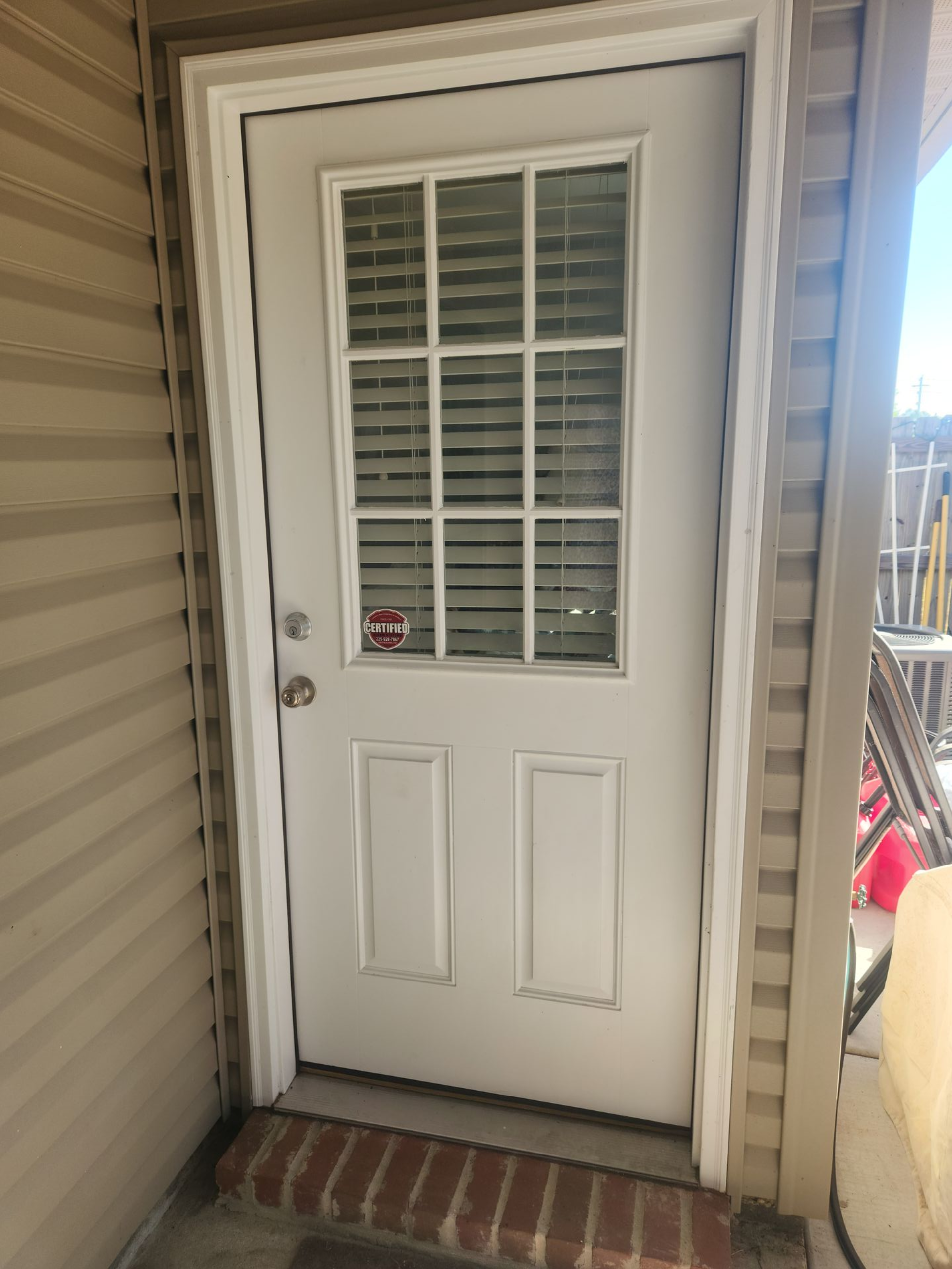 White exterior door with window panes, brick step, and beige siding beside a narrow porch area