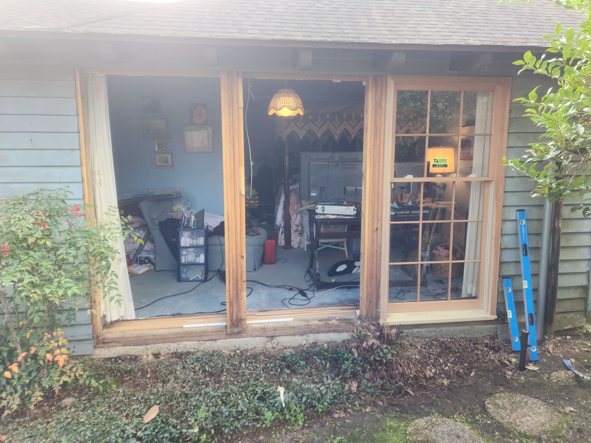 Open double doors and windows of a house, with a cluttered interior and blue ladder outside.