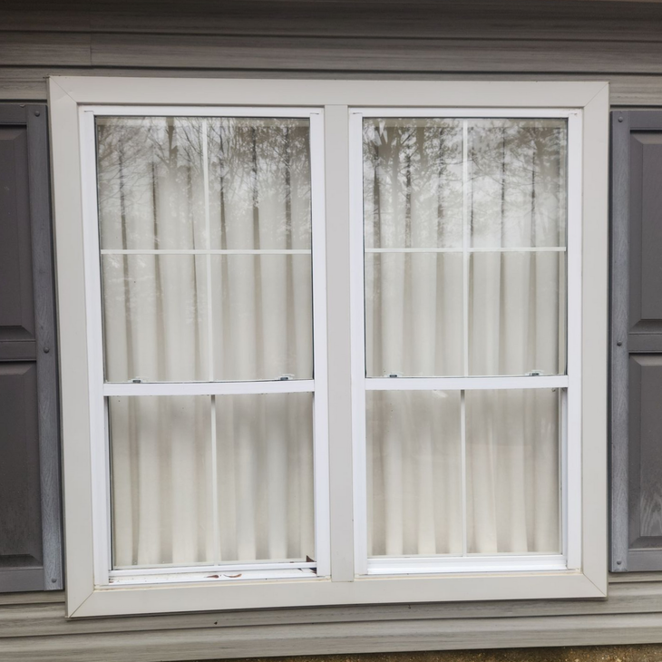 White-framed double window with closed blinds on a gray house exterior.