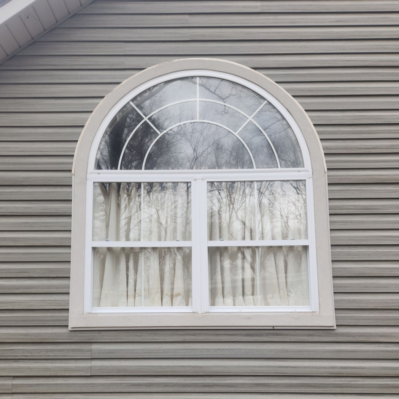 Arched window with white trim and curtains set in gray vinyl siding.