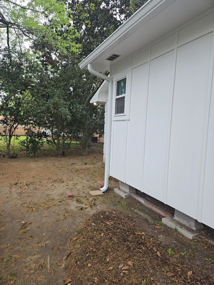 Side of a white house with a small window, downspout, and leaf-covered yard beside trees.