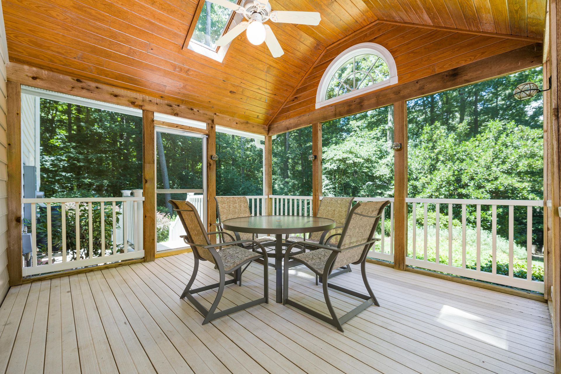 Covered wooden porch with ceiling fan, railing, and patio table and chairs overlooking trees