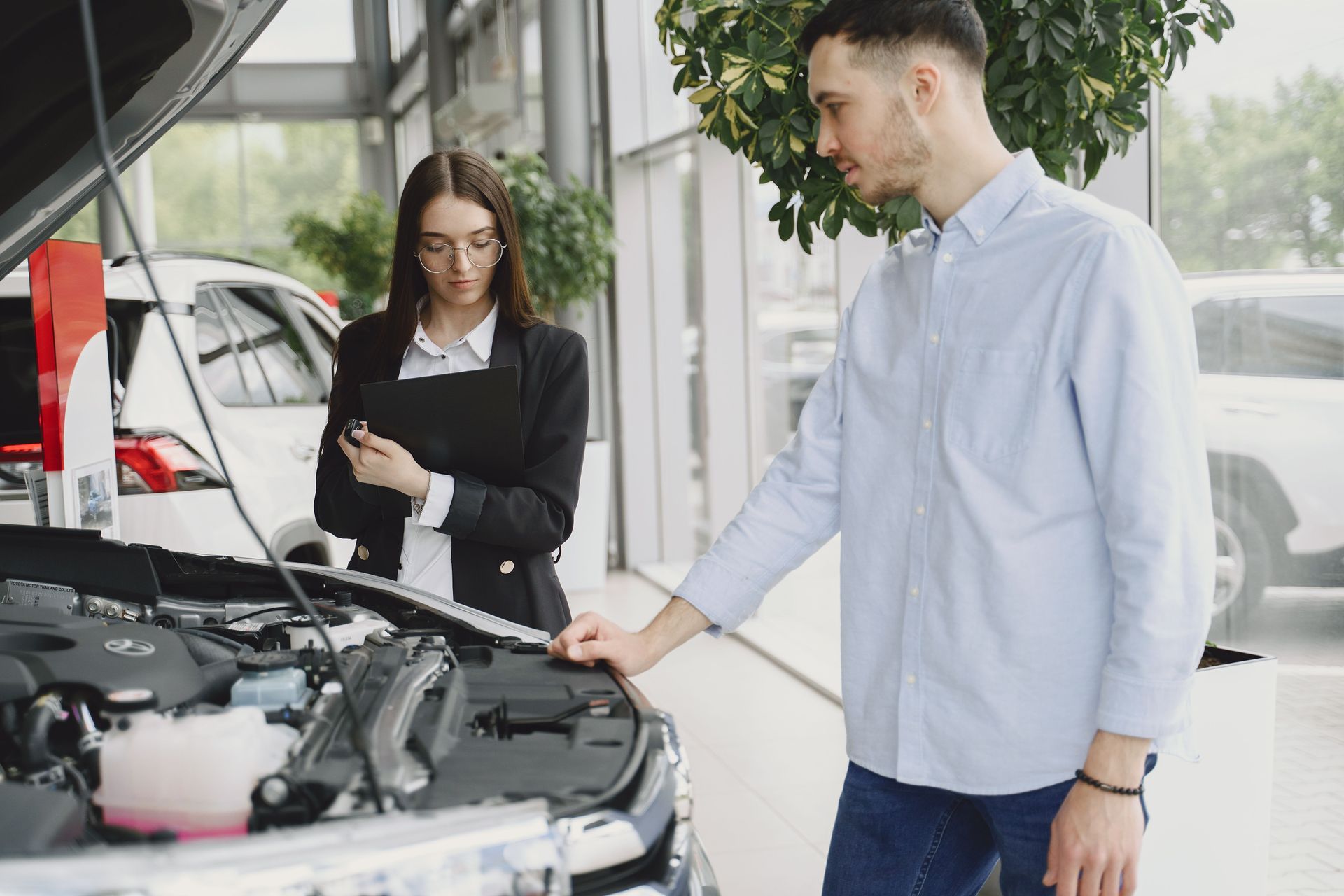 Person and woman inspecting an open car engine in a bright auto showroom